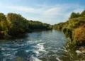 A serene river flows between green, tree-lined banks under a blue sky with light clouds.