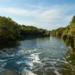 A serene river flows between green, tree-lined banks under a blue sky with light clouds.