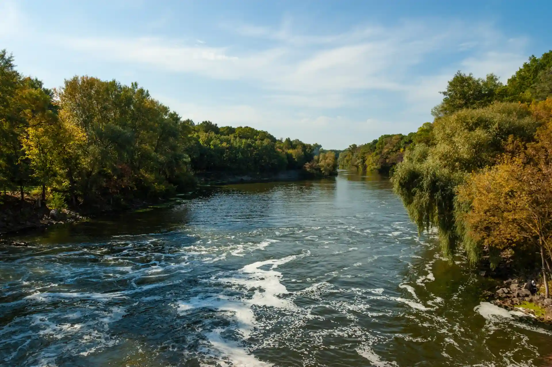 A serene river flows between green, tree-lined banks under a blue sky with light clouds.