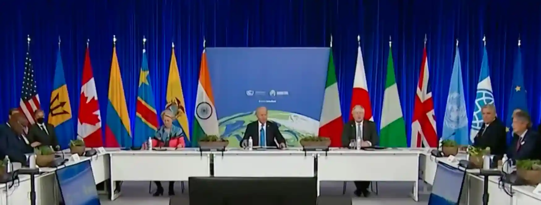 World leaders sit at a large conference table on a stage with flags of various countries in the background, during an international summit with blue curtains. The discussion prominently features the Renewable energy transition and key COP summit outcomes.