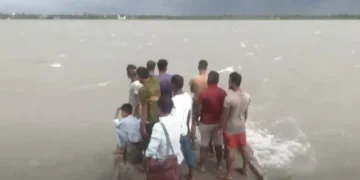 A group of people stands on a small boat in a large body of water under a cloudy sky, likely navigating the aftermath of Cyclone Remal near Bangladesh.