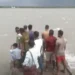 A group of people stands on a small boat in a large body of water under a cloudy sky, likely navigating the aftermath of Cyclone Remal near Bangladesh.