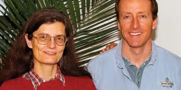 A woman and a man stand together in front of a green plant. The woman wears glasses, a red sweater, and a checked shirt. The man, who has been with Harrington's Organic Land Care for 35 years, wears a blue button-down shirt with a logo on it and smiles proudly.