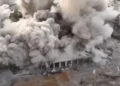 Aerial view of a large building collapsing amidst thick plumes of smoke and debris, with nearby structures partially visible, a stark representation of the ongoing Gaza conflict.