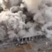 Aerial view of a large building collapsing amidst thick plumes of smoke and debris, with nearby structures partially visible, a stark representation of the ongoing Gaza conflict.