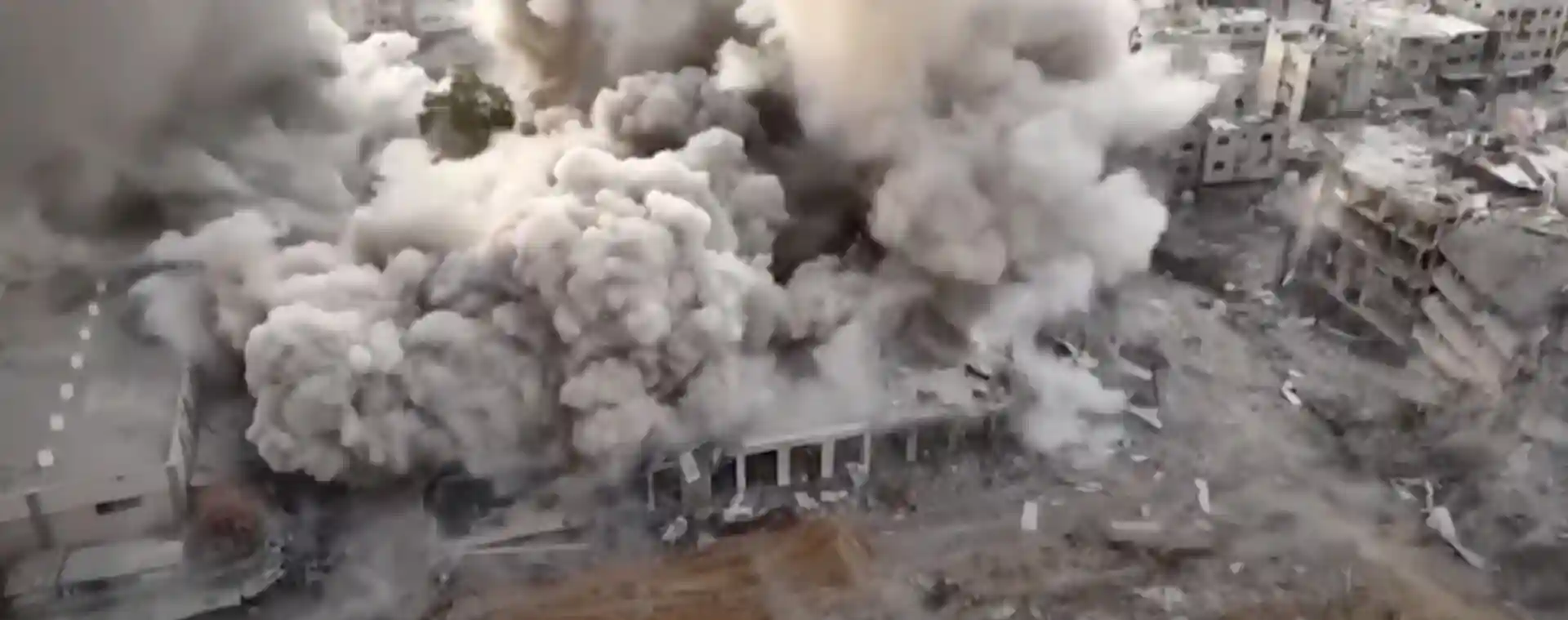 Aerial view of a large building collapsing amidst thick plumes of smoke and debris, with nearby structures partially visible, a stark representation of the ongoing Gaza conflict.
