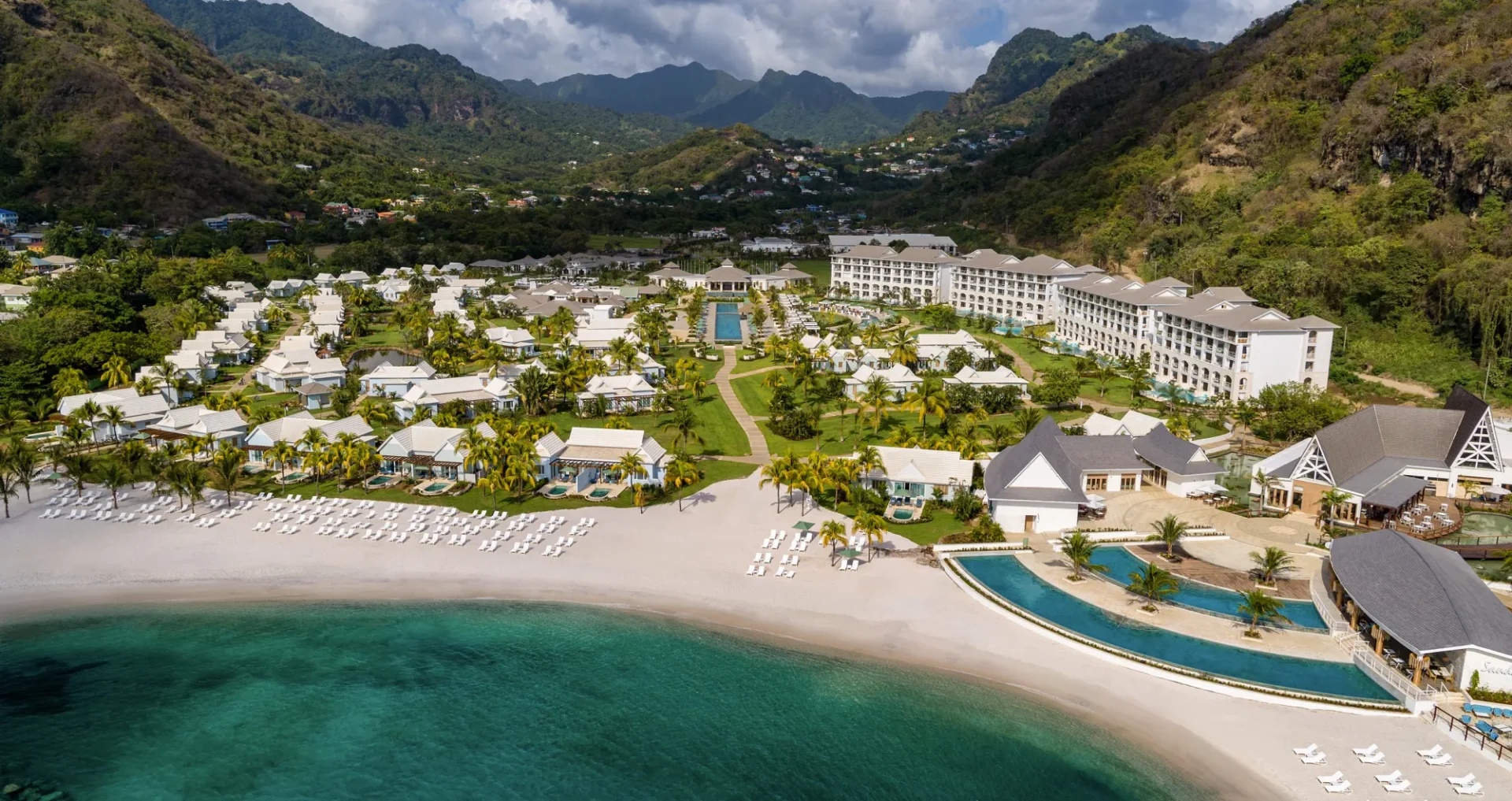 An aerial view of a beachfront resort in St. Vincent and the Grenadines, showcasing multiple buildings, a pool, and sandy beach surrounded by lush, green mountains and turquoise water—a true gem of Caribbean luxury travel.