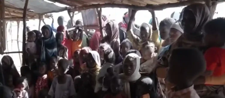 A large group of people, including children and adults, gather inside a wooden shelter with a thatched roof. Many are wearing headscarves and looking toward the camera, reflecting the ongoing Sudan crisis 2024.