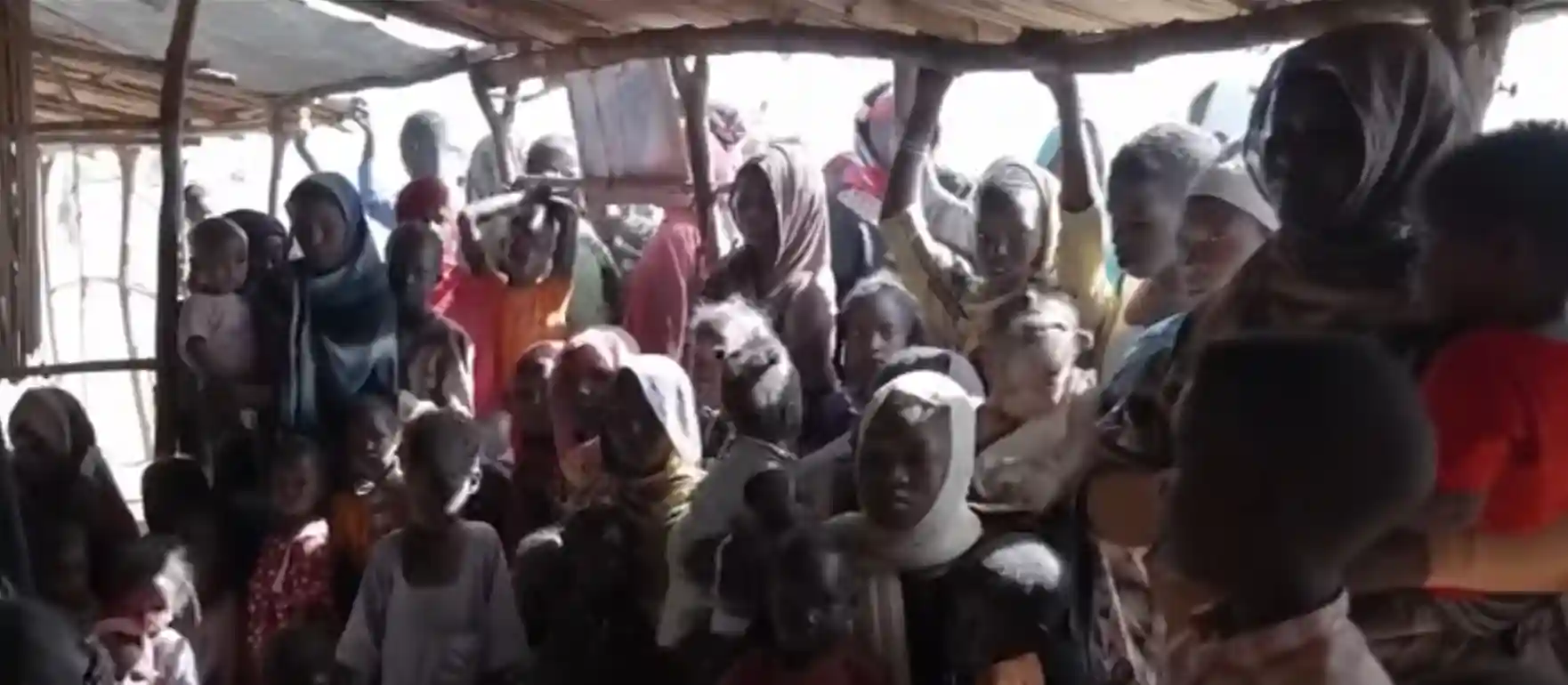 A large group of people, including children and adults, gather inside a wooden shelter with a thatched roof. Many are wearing headscarves and looking toward the camera, reflecting the ongoing Sudan crisis 2024.
