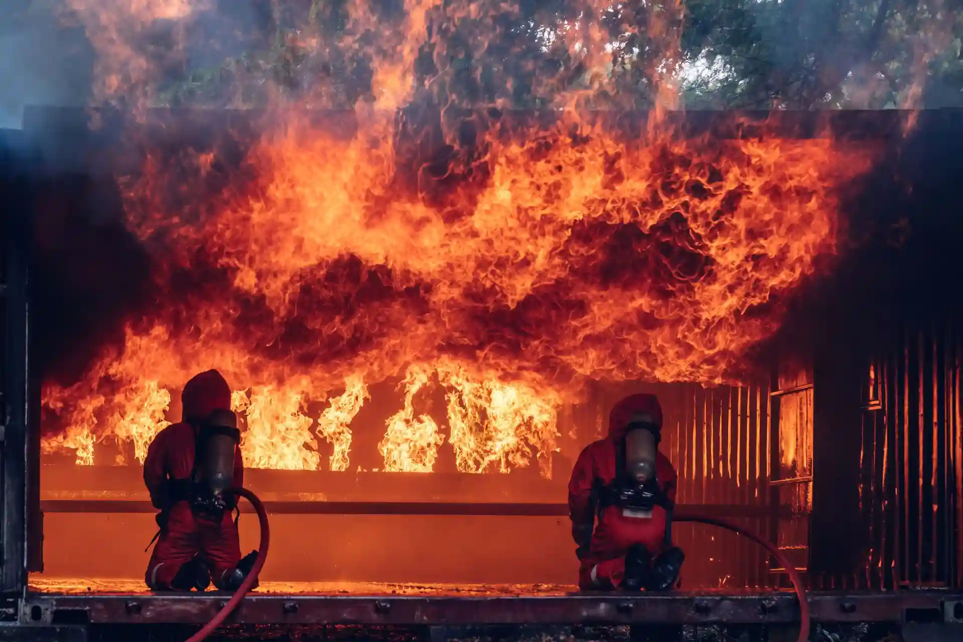 Two firefighters in full gear kneel in front of a large, intense blaze inside a structure, aiming their hoses to combat the fire.