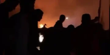 Silhouetted figures stand against a backdrop of orange flames and heavy smoke at night, reminiscent of scenes from Rafah following recent Israeli airstrikes.