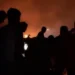 Silhouetted figures stand against a backdrop of orange flames and heavy smoke at night, reminiscent of scenes from Rafah following recent Israeli airstrikes.