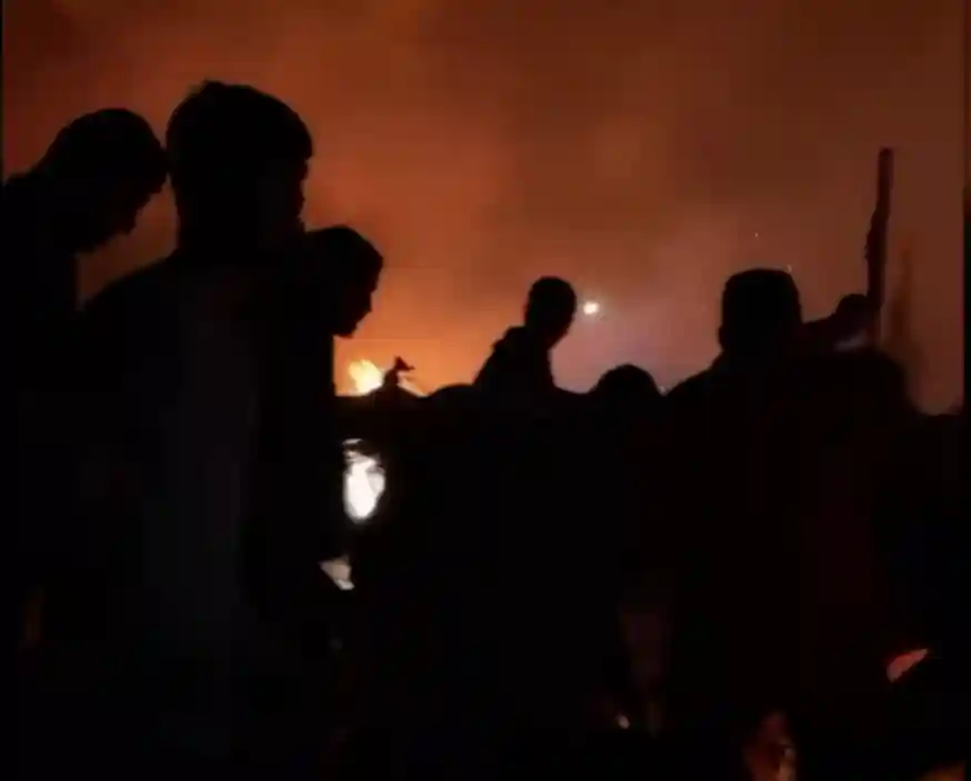 Silhouetted figures stand against a backdrop of orange flames and heavy smoke at night, reminiscent of scenes from Rafah following recent Israeli airstrikes.