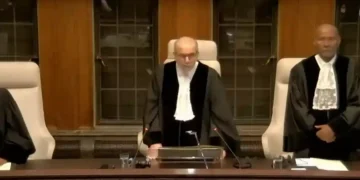 Judges dressed in formal judicial gowns stand in a courtroom, one is speaking into a microphone while others stand on either side, discussing the ramifications of the Rafah Offensive.