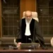 Judges dressed in formal judicial gowns stand in a courtroom, one is speaking into a microphone while others stand on either side, discussing the ramifications of the Rafah Offensive.