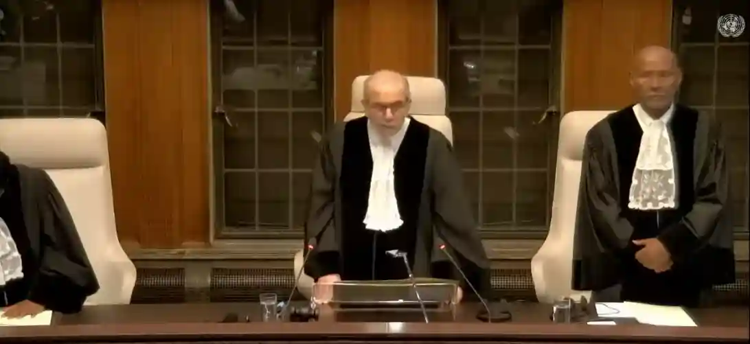 Judges dressed in formal judicial gowns stand in a courtroom, one is speaking into a microphone while others stand on either side, discussing the ramifications of the Rafah Offensive.