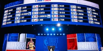 A large LED screen shows the order of NBA draft picks for 2024. Below the screen, a podium is set up under bright lights, with "DRAFT 2024" and NBA logos prominently displayed, making it the centerpiece for all 2024 NBA Draft news.