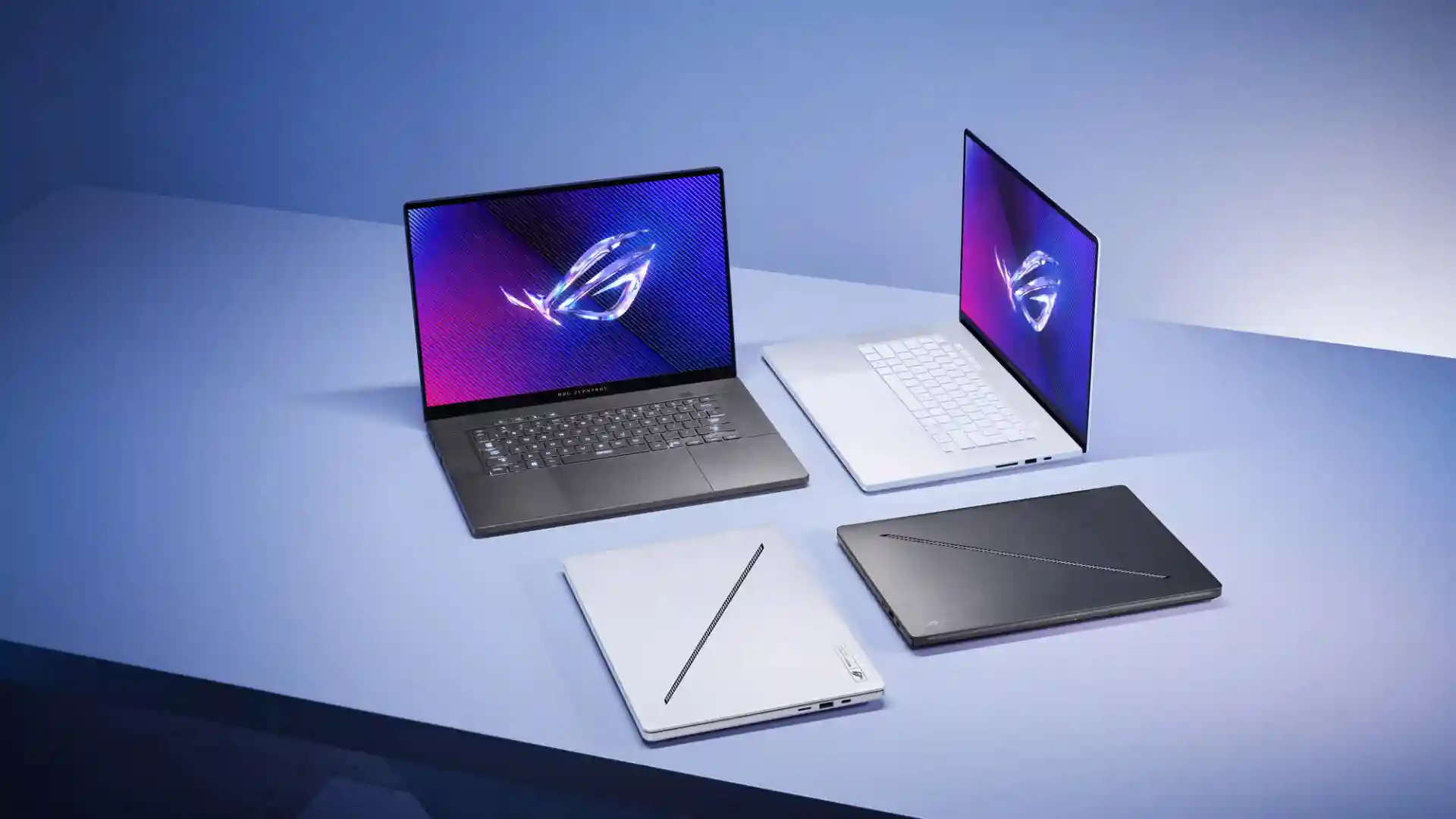 A collection of four laptops on a light blue surface. Two are open, one facing forward and the other at an angle, displaying colorful screens with a logo. Among them is the high-end gaming ASUS ROG Zephyrus G16. Two laptops are closed, one white and one black.
