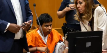A person in an orange jumpsuit sits in a courtroom with a bandaged arm, surrounded by NYPD officers, legal professionals, and law enforcement.