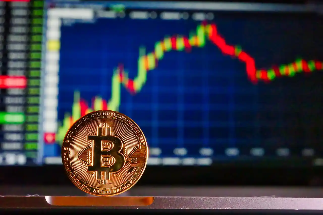 A Bitcoin coin in the foreground with a cryptocurrency price chart displaying fluctuating values on a screen in the background, showcasing BTC analysis trends.