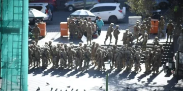 A group of uniformed soldiers gather in a city's public square near parked cars and orange barriers, amid rising tensions from a recent coup attempt. Pigeons are visible in the foreground.