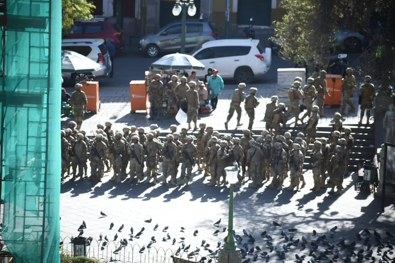 A group of uniformed soldiers gather in a city's public square near parked cars and orange barriers, amid rising tensions from a recent coup attempt. Pigeons are visible in the foreground.