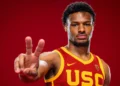 A basketball player in a USC jersey, potentially Bronny James, holds up two fingers against a red background as he eyes spots in the 2024 NBA Draft.
