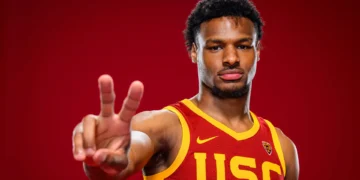 A basketball player in a USC jersey, potentially Bronny James, holds up two fingers against a red background as he eyes spots in the 2024 NBA Draft.