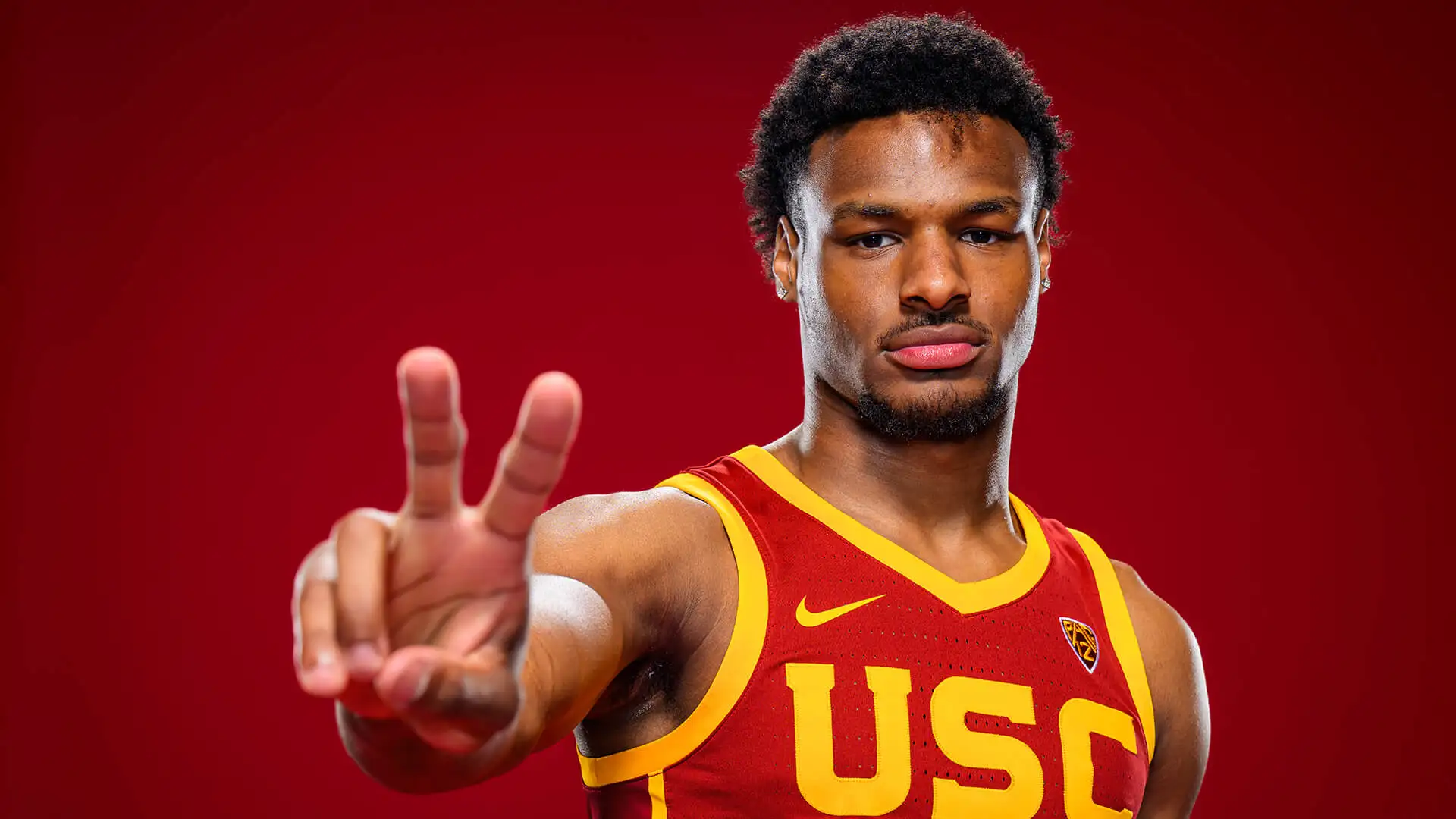 A basketball player in a USC jersey, potentially Bronny James, holds up two fingers against a red background as he eyes spots in the 2024 NBA Draft.