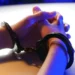 Close-up of a person's hands in handcuffs, with fingers interlaced, resting on a table under blue and white lighting. The image evokes scenes often seen in gang-related videos posted by a popular Canadian vlogger.