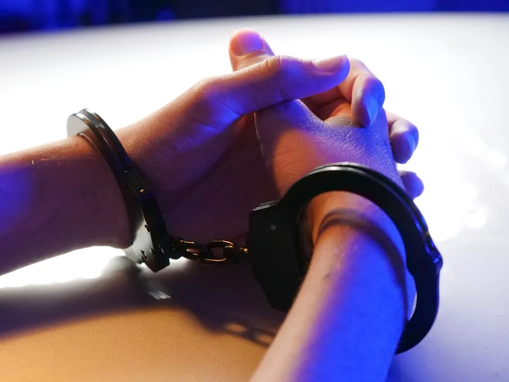 Close-up of a person's hands in handcuffs, with fingers interlaced, resting on a table under blue and white lighting. The image evokes scenes often seen in gang-related videos posted by a popular Canadian vlogger.