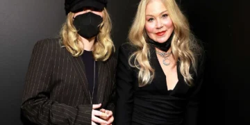 Two women with long blonde hair stand next to each other. One wears a black hat and face mask, while the other, who resembles Christina Applegate, is dressed in black with several necklaces. Both are smiling, seemingly carefree despite one’s daughter diagnosed with POTS.