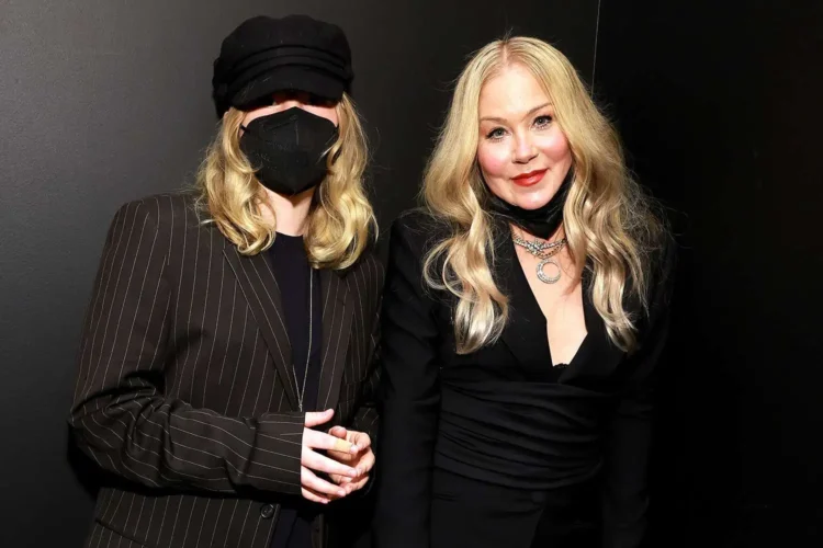 Two women with long blonde hair stand next to each other. One wears a black hat and face mask, while the other, who resembles Christina Applegate, is dressed in black with several necklaces. Both are smiling, seemingly carefree despite one’s daughter diagnosed with POTS.
