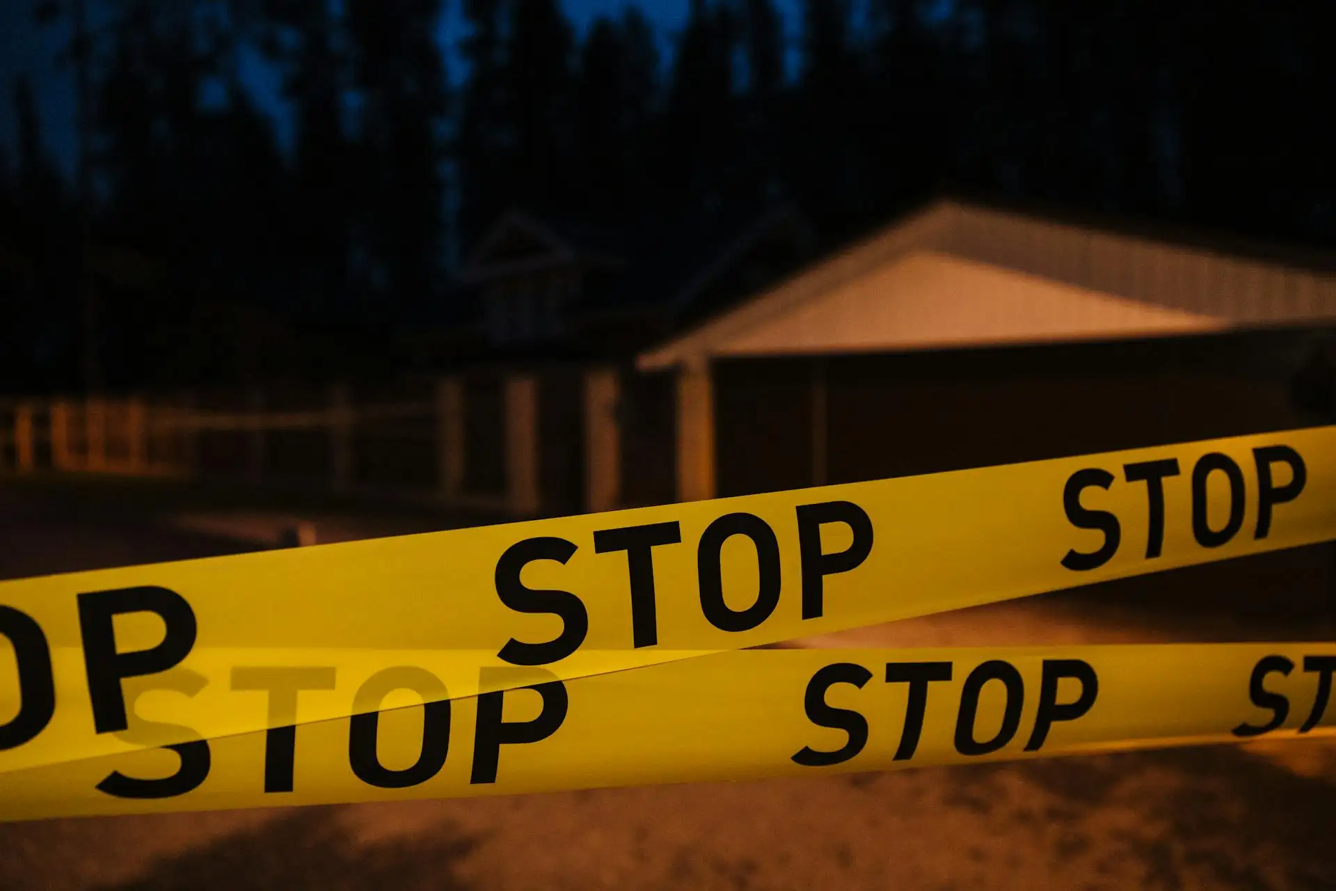 Yellow "STOP" tape cordons off an area in front of a house at night, casting a somber pall over the neighborhood as trees and other structures loom in the background—marking the site of a recent oven tragedy in Kansas City.