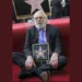 An older man with white hair and beard, dressed in a suit, sits on red carpet holding a plaque of a star. The iconic actor Donald Sutherland proudly showcases his well-deserved accolade.