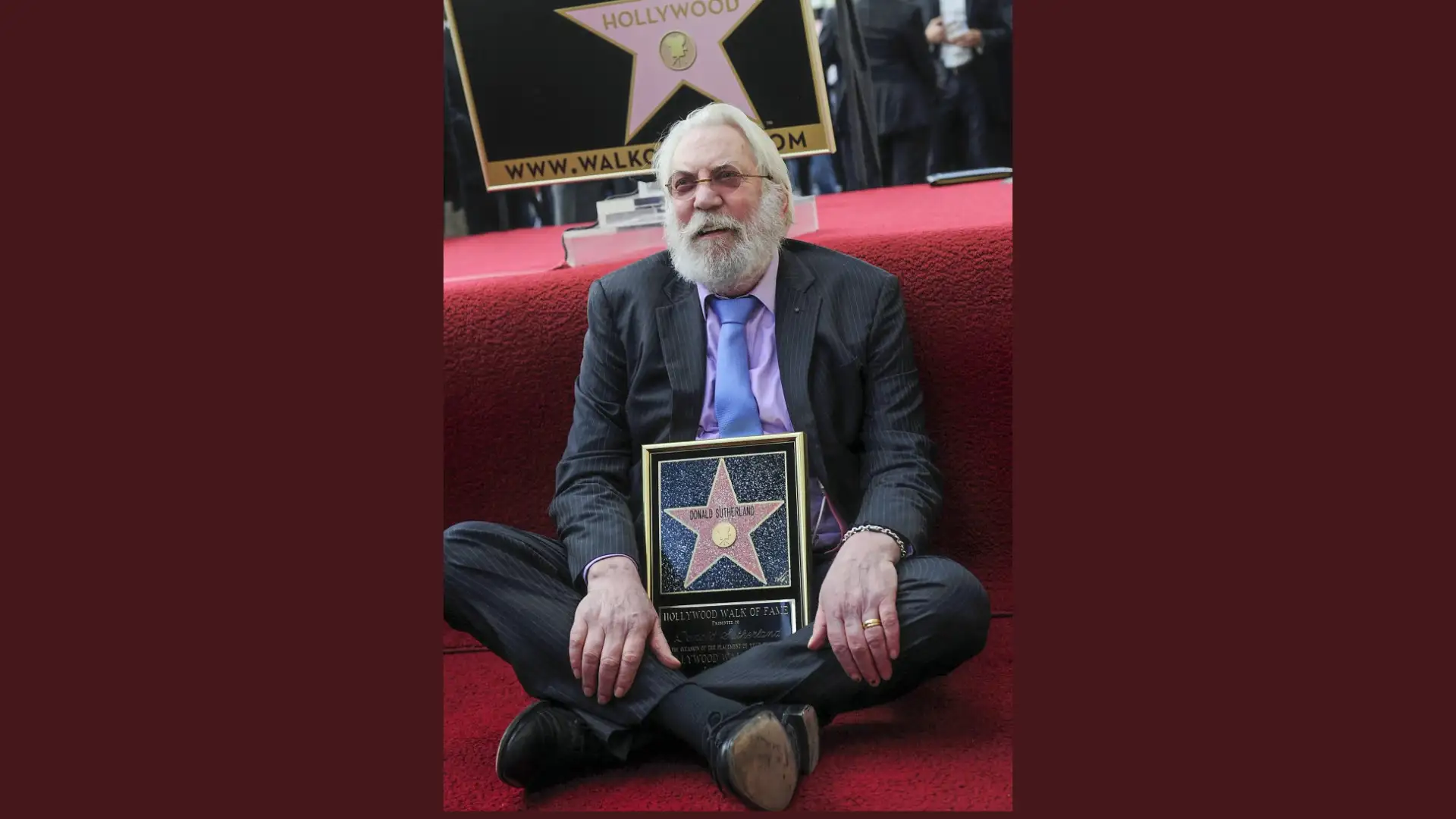 An older man with white hair and beard, dressed in a suit, sits on red carpet holding a plaque of a star. The iconic actor Donald Sutherland proudly showcases his well-deserved accolade.