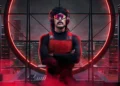 A person with a mustache and sunglasses, wearing a red vest and headphones, stands with arms crossed against a backdrop of a circular window with a city skyline illuminated in red. It’s like Dr. Disrespect after being featured in the latest gaming news, post-Twitch ban.