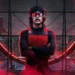 A person with a mustache and sunglasses, wearing a red vest and headphones, stands with arms crossed against a backdrop of a circular window with a city skyline illuminated in red. It’s like Dr. Disrespect after being featured in the latest gaming news, post-Twitch ban.