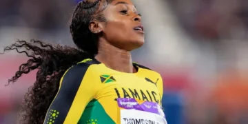 Elaine Thompson-Herah in a green, yellow, and black uniform, injured after competing in a sprinting event at the New York Grand Prix, with a visible Jamaican flag and 'Jamaica' written on her outfit.