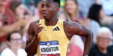 A male athlete, Erriyon Knighton, wearing a yellow racing outfit with the name "Knighton" and number "1" displayed on his chest, runs on a track during a race as part of the U.S. Olympic Team, achieving an impressive 19.77-second finish.