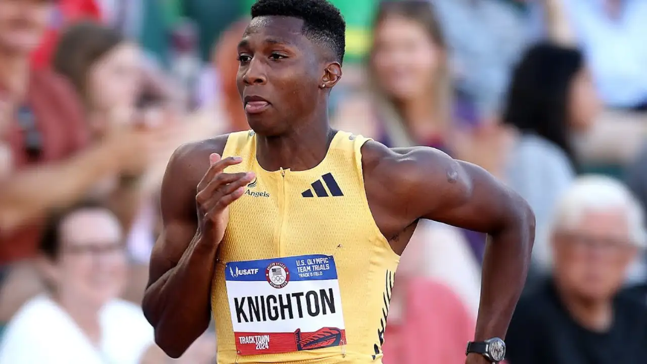 A male athlete, Erriyon Knighton, wearing a yellow racing outfit with the name "Knighton" and number "1" displayed on his chest, runs on a track during a race as part of the U.S. Olympic Team, achieving an impressive 19.77-second finish.