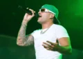 A person wearing a green cap, sunglasses, and a white t-shirt is singing into a microphone on stage under green lighting, reminiscent of Feid's energetic performances that might be seen at the Copa América 2024 in Atlanta.