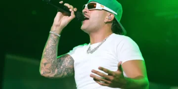 A person wearing a green cap, sunglasses, and a white t-shirt is singing into a microphone on stage under green lighting, reminiscent of Feid's energetic performances that might be seen at the Copa América 2024 in Atlanta.