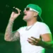 A person wearing a green cap, sunglasses, and a white t-shirt is singing into a microphone on stage under green lighting, reminiscent of Feid's energetic performances that might be seen at the Copa América 2024 in Atlanta.