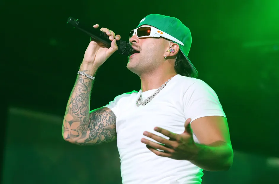 A person wearing a green cap, sunglasses, and a white t-shirt is singing into a microphone on stage under green lighting, reminiscent of Feid's energetic performances that might be seen at the Copa América 2024 in Atlanta.