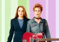 Two women stand against a pastel-striped background. One, reminiscent of Lindsay Lohan's character in Freaky Friday 2, is in formal attire with pearls, while the other channels Disney spirit in a denim jacket with short spiky hair, holding a red electric guitar.