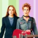Two women stand against a pastel-striped background. One, reminiscent of Lindsay Lohan's character in Freaky Friday 2, is in formal attire with pearls, while the other channels Disney spirit in a denim jacket with short spiky hair, holding a red electric guitar.