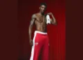 A shirtless man with a muscular build stands confidently in front of a red curtain, holding a white towel. He is wearing red pants with white stripes and Olympic rings on the waist.