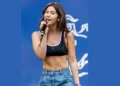 A woman with long dark hair is singing into a microphone on stage, wearing a black crop top and blue jeans, with a blue background behind her. Her performance exudes the excitement of new music, reminiscent of the energy found on a Taylor Swift tour.