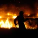 A firefighter silhouetted against a raging fire at night, holding a hose and navigating through grass as flames blaze in the background.